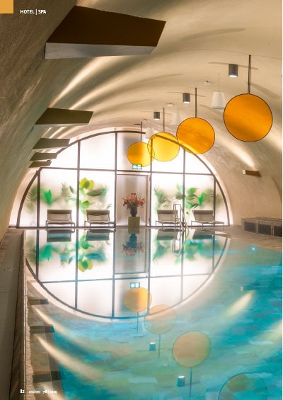 Modernes Spa mit geschwungenem Dach, gelben Lichtern und Wandkunst, reflektiert im Wasser.